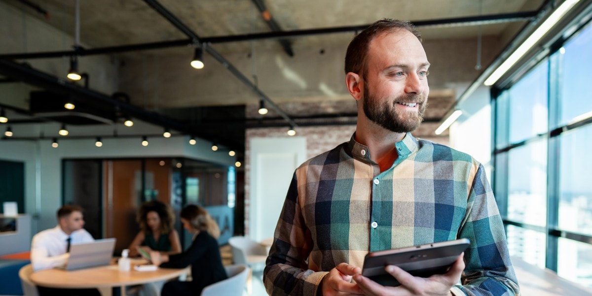 man in the office looking out window, holding tablet, colleagues sitting at a table in the background