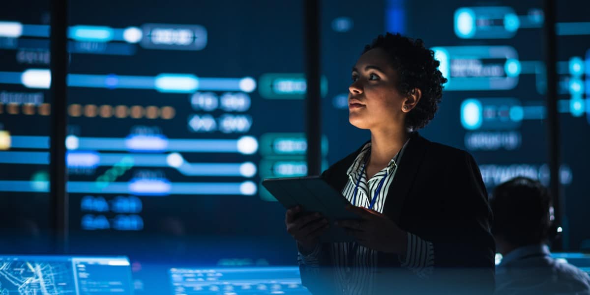 professional woman in an office, holding tablet, lit up screens around her showing data graphics
