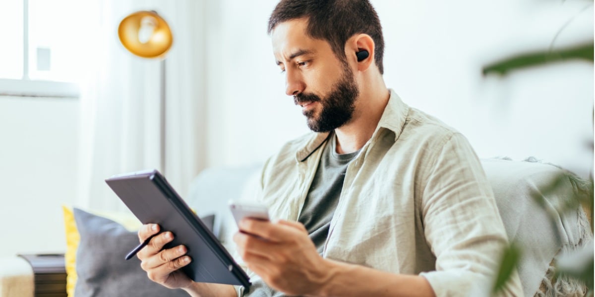 young bearded man working from home on a sofa with a tablet and headphones